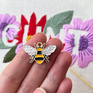 May include: A gold-colored bee charm with black and yellow stripes and white wings. The charm is held in a hand, with a blurred background of embroidered flowers in red and purple.