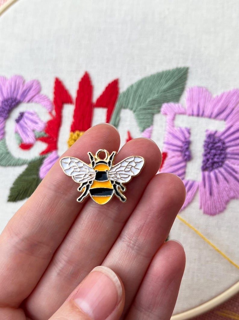 May include: A gold-colored bee charm with black and yellow stripes and white wings. The charm is held in a hand, with a blurred background of embroidered flowers in red and purple.