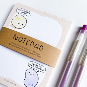 May include: A notepad with a brown cover and white pages. The cover has a cartoon illustration of a cell with the text "you're LEUKIN great!" and "C3". The back of the notepad has a cartoon illustration of a cell with the text "thanks for the COMPLEMENT!" and "IL-6". The notepad is next to two purple pens.