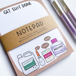 May include: A notepad with the text "GET SHIT DONE" at the top. The notepad has a brown band that reads "NOTEPAD" and includes the website and social media handle. The bottom of the notepad features cartoon illustrations of medicine bottles and a container. Two purple pens are on the right.