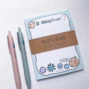 May include: A notepad with the text "B Adaptive!" and "NOTEPAD" is shown, along with two pens. The notepad has a light blue border and a brown band. The pens are pink and teal.
