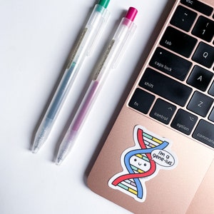 Op de afbeelding: Twee transparante pennen met groene en roze dopjes op een roze gouden laptop met een sticker van een cartoon DNA-streng met een spraakballon die "i'm a gene-ius" zegt.