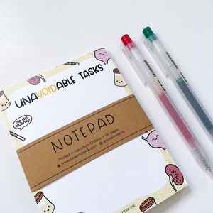 May include: A white notepad with a yellow background and a brown paper band. The notepad has a cartoon design with illustrations of a jar of milk, a kidney, and a speech bubble that says "dip dip hooray!" The band around the notepad says "UNAVOIDABLE TASKS NOTEPAD Printed in Hamilton, Ontario 50 pages www.soapnoteink.com @soapnoteink".