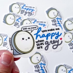 May include: A white sticker with a cartoon face and the text "SAD no more!". The sticker also says "you make me happy when skies are grey" in blue and grey script. A small red heart is included in the design. The sticker is held by a hand.