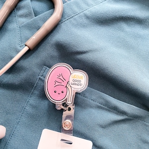 May include: A pink cartoon kidney with a smiling face and the text "Urine Good Hands!" on a white badge holder. The badge holder is attached to a teal scrub top.