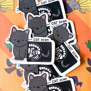 May include: A stack of black and white cat scan stickers with a cartoon cat on each sticker. The stickers say "CAT scan" and "soap note ink".