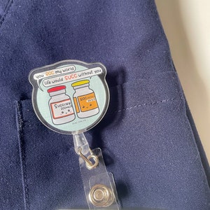 May include: A blue scrub top with a clear plastic badge holder attached. The badge holder has a round sticker with a cartoon image of two medicine bottles. The sticker says "you ROC my world life would SUCC without you".