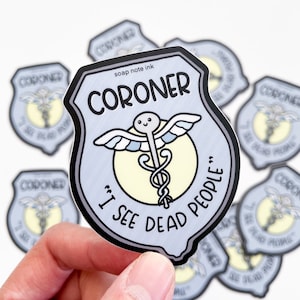May include: A light blue sticker with a shield design featuring the word "CORONER" and the phrase "I SEE DEAD PEOPLE." The sticker includes a caduceus symbol and a smiling face. Several stickers are visible.