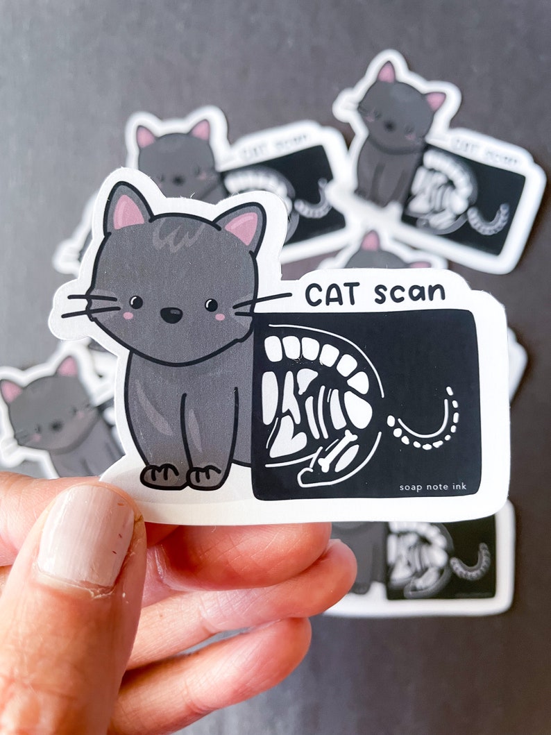 May include: A gray cat sticker with a cartoon illustration of a cat and an x-ray image of a cat skeleton. The sticker says "CAT scan" and "soap note ink".