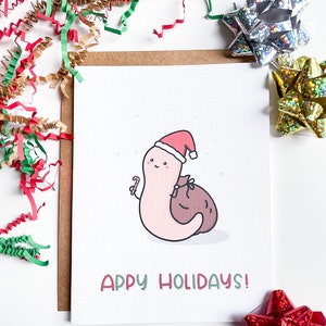 May include: A white card with a cartoon worm wearing a Santa hat and holding a candy cane. The worm is carrying a brown sack. The card says "Happy Holidays!" in green and red letters.
