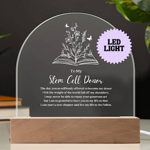 May include: A white acrylic plaque with a wood base and a LED light. The plaque features a floral design with a book and the text "To My Stem Cell Donor". The text reads: "The day you so selflessly offered to become my donor I felt the weight of the world fall off my shoulders. I may never be able to repay your generous act but I am so grateful to have you in my life so that I can start a new chapter and live my life to the fullest."
