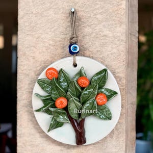 May include: A white ceramic wall hanging featuring a green and orange citrus tree design. The tree has a brown trunk and is adorned with a blue evil eye charm.