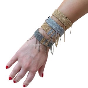 May include: Several gold and silver-toned chain bracelets with dangling fringe are displayed on a wrist. The bracelets are woven in a wide band style. The hand is positioned against a white background.