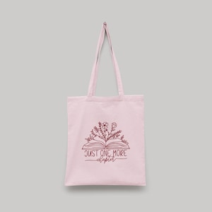 Peut inclure: Un sac fourre-tout rose clair avec un motif de livre et de fleurs. Le sac a une illustration marron d'un livre ouvert avec des fleurs et le texte "JUST ONE MORE chapter". Le sac a deux longues anses.
