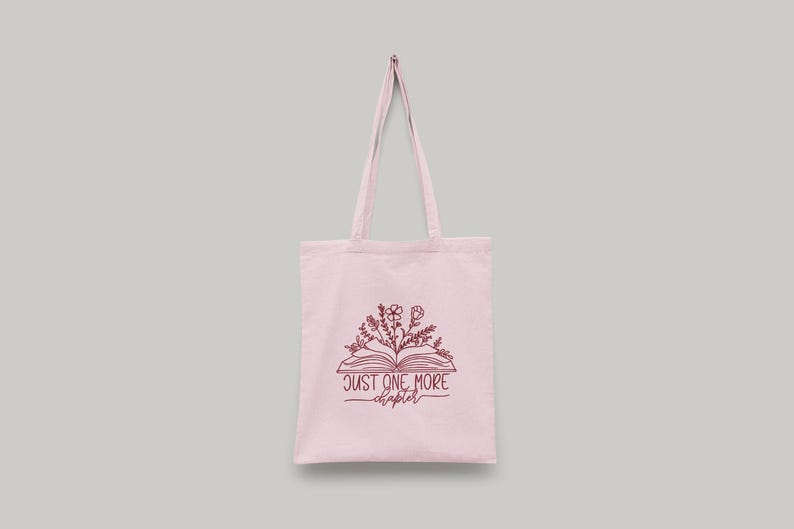 Peut inclure: Un sac fourre-tout rose clair avec un motif de livre et de fleurs. Le sac a une illustration marron d'un livre ouvert avec des fleurs et le texte "JUST ONE MORE chapter". Le sac a deux longues anses.