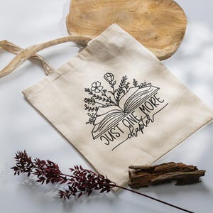 Peut inclure: Sac fourre-tout en toile beige avec un dessin au trait noir d'un livre ouvert, de fleurs et le texte "JUST ONE MORE chapter". Le sac a de longues anses et est pos&eacute; sur une surface blanche avec des &eacute;l&eacute;ments d&eacute;coratifs.
