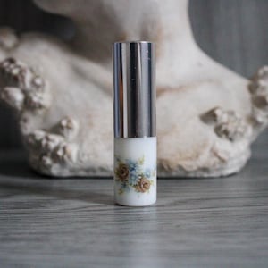 Op de afbeelding: Een kleine, cilindrische parfumfles met een zilveren dop en een witte basis. De basis is versierd met een bloemmotief in blauw- en bruintinten. De fles staat rechtop.
