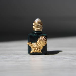 Peut inclure: Petite bouteille de parfum vert foncé avec un motif d'oiseau doré et un bouchon doré. Le bouchon est surmonté d'une petite perle blanche. La bouteille est rectangulaire et a une surface texturée.