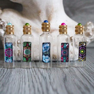 Op de afbeelding: Vijf heldere glazen flessen met decoratieve etiketten en goudkleurige doppen. Elke fles heeft een ander kleurrijk ontwerp op het etiket. De doppen zijn bekroond met een kleine, gekleurde edelsteen. De flessen zijn gerangschikt op een houten oppervlak.
