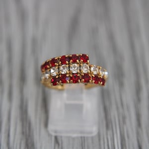 Può includere: Un anello dorato con tre file di pietre preziose rosse e bianche. L'anello è esposto su un supporto trasparente su uno sfondo grigio sfocato. Le pietre preziose sono tagliate a forma quadrata e disposte in un motivo ripetuto.