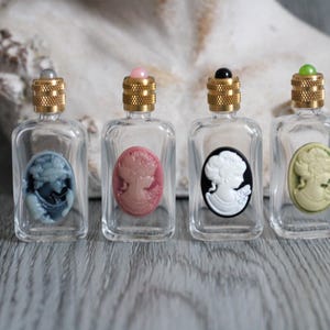 Op de afbeelding: Vier kleine, heldere glazen parfumflesjes met goudkleurige deksels en camee-ontwerpen. De cameeën zijn in tinten blauw, roze, zwart en wit en groen. Elke fles heeft een verschillend gekleurde edelsteen bovenop.