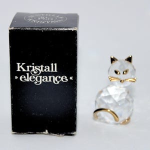 May include: A clear crystal cat figurine with gold accents, sitting next to a black box with the text "Kristall elegance". The cat has a faceted design and gold-colored ears and whiskers.