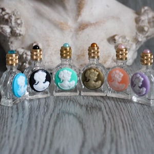 May include: Six small, clear glass perfume bottles with gold-colored tops and cameo designs in various colors. The cameo colors include blue, black, green, brown, orange, and purple. The bottles are arranged in a row.