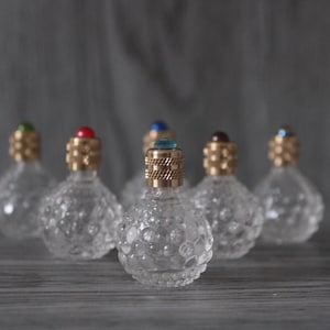 Peut inclure: Plusieurs petits flacons de parfum en verre transparent avec des corps ronds texturés. Chaque flacon est surmonté d'un bouchon doré et d'une gemme colorée, rouge, bleue et verte. Les flacons sont disposés sur une surface grise.