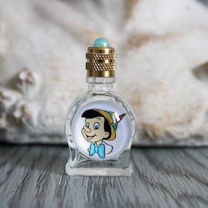 May include: A small, clear glass perfume bottle with a gold-colored cap and a light blue gem on top. The bottle features a circular image of Pinocchio. The bottle is sitting on a gray wooden surface.