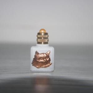 Könnte beinhalten: Kleine, weiße Parfümflasche mit einem kupferfarbenen Katzenkopf-Emblem. Die Flasche hat einen goldfarbenen Verschluss und einen kleinen, runden, bernsteinfarbenen Stopfen. Die Flasche ist rechteckig mit abgerundeten Kanten.