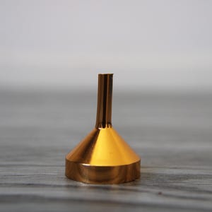 May include: A gold-coloured metal funnel with a wide, conical base and a narrow, cylindrical spout. The funnel is made of a shiny, reflective material, and it is designed to pour liquids or fine-grained substances into containers. The funnel is approximately 10 cm tall.