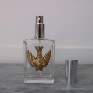 May include: A clear, rectangular perfume bottle with a gold-colored bird emblem. The bottle has a silver-colored spray nozzle and a separate silver cap. The bird is depicted with outstretched wings. The bottle is on a gray surface.