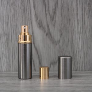 Op de afbeelding: Een zilverkleurige parfumverstuiver met een goudkleurige bovenkant en bijpassende dop. De verstuiver is cilindrisch, met een sproeikop. Er worden twee doppen getoond, één goudkleurig en één zilverkleurig. De achtergrond is een grijze houtnerf.