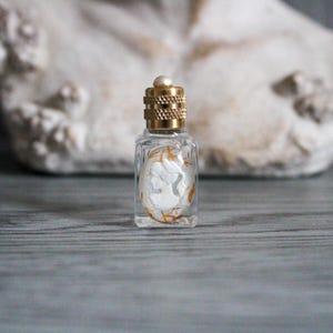 May include: A small, clear glass perfume bottle with a gold-colored cap and a pearl on top. Inside the bottle is a white cameo with a profile image. The cameo is surrounded by gold and orange accents.