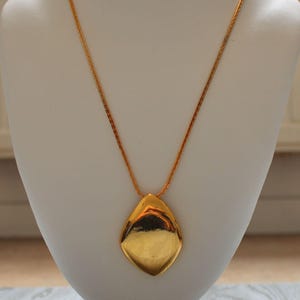 May include: A gold-toned necklace with a large, teardrop-shaped pendant. The pendant has a smooth, reflective surface. The chain is delicate and also gold-toned. The necklace is displayed on a white bust.