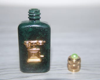 May include: A dark green, rectangular bottle with a gold-colored rotary phone design. The bottle has a matching gold-colored cap with a green gem on top. The bottle is made of a speckled, marble-like material.