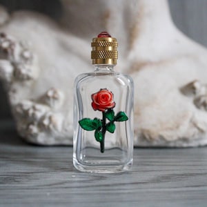 Könnte beinhalten: Eine transparente Glasparfümflasche mit einem roten Rosendesign im Inneren. Die Flasche hat einen goldfarbenen Deckel mit einem roten Edelstein. Die Rose hat grüne Blätter und einen schwarzen Stiel. Die Flasche ist rechteckig.