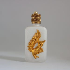 Op de afbeelding: Een witte, rechthoekige parfumfles met een goudkleurig eekhoornontwerp. De fles heeft een goudkleurige, getextureerde dop met een kleine parel. De eekhoorn staat in een speelse pose.