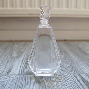 Peut inclure: Flacon de parfum clair de style Art déco avec un bouchon géométrique. La bouteille a une forme évasée en forme d'éventail et repose sur une surface en bois. Le bouchon est une série de formes claires et angulaires.