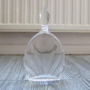 Peut inclure: Une bouteille de parfum en verre transparent avec un bouchon facetté. La bouteille a une base arrondie et un motif en forme d'éventail sur le devant. Le bouchon est de forme claire et pointue. La bouteille repose sur une surface en bois grise.