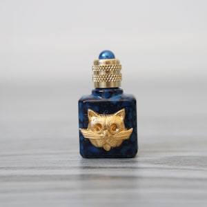 Op de afbeelding: Een kleine, vierkante, donkerblauwe fles met een gouden kattengezicht-embleem. De fles heeft een goudkleurige, getextureerde dop en een blauwe edelsteen bovenop. Het kattengezicht heeft gedetailleerde kenmerken, waaronder snorharen en grote ogen.