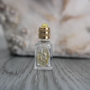 Può includere: Piccola bottiglia di profumo in vetro trasparente con un tappo dorato strutturato e una perla gialla sulla parte superiore. La bottiglia contiene un elemento decorativo dorato. La bottiglia è esposta su una superficie di legno grigio.