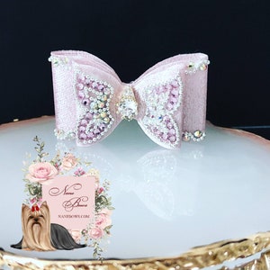 Può includere: Un fiocco rosa glitterato con un design a farfalla e strass. Il fiocco è su una superficie bianca con un bordo dorato.