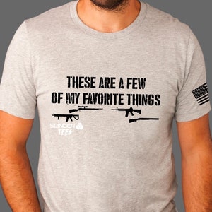Puede incluir: Una camiseta gris con el texto "These are a few of my favorite things" e imágenes de rifles. La camiseta también tiene una pequeña bandera estadounidense en la manga y el texto "Slunger Tees" en la parte inferior.