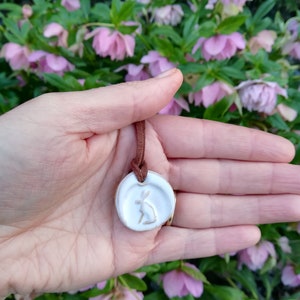 May include: A white ceramic pendant with a brown leather cord. The pendant features a raised design of a rabbit with a leaf in its mouth.