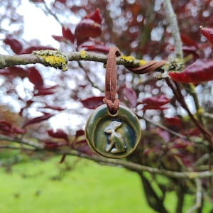 Op de afbeelding: Een ronde, groene keramische hanger met een reliëf van een konijn, bevestigd aan een bruin leren koord. De hanger hangt aan een tak met rode bladeren en groen gras op de achtergrond.