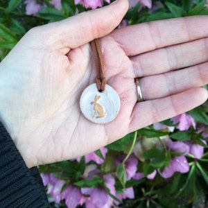 May include: A white ceramic pendant featuring a gold-coloured silhouette of a rabbit, suspended from a brown leather cord. The pendant is being held in a hand.