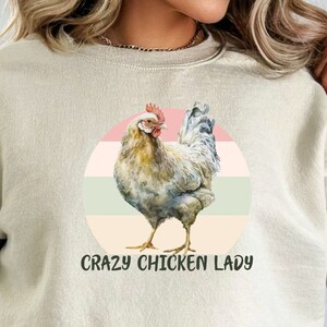 May include: A beige sweatshirt with a graphic of a white chicken in a circle with pink, green, and white stripes. The text "Crazy Chicken Lady" is printed below the graphic.
