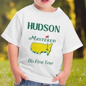 Könnte beinhalten: Weißes T-Shirt mit dem Namen "HUDSON" in grünen Buchstaben, über den Wörtern "MASTERED" und "His First Year". Eine gelbe Kartengrafik mit einer Golf-Flagge ist auf dem T-Shirt zentriert. Das T-Shirt wird von einem Kind getragen.