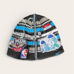 May include: A striped knitted beanie hat in grey, blue, and white, decorated with patches including a New Orleans Pelicans logo, a Nike logo, and a Greek-style building. Features a small Nike swoosh and a decorative string.